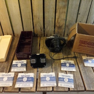 May include: A vintage View-Master projector and a collection of View-Master reels. The projector is black and has a round lens. The reels are in their original packaging and feature images of the world. The projector is in its original box, which is brown and has the words "View-Master Junior Projector" printed on it.