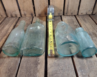 May include: Four vintage glass bottles with a green tint. The bottles are various sizes and shapes. The bottles are sitting on a wooden surface.