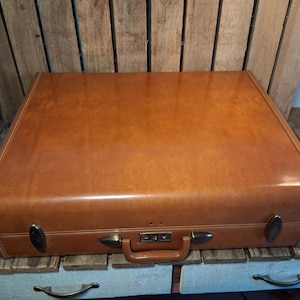May include: A rectangular, tan-colored hard-shell suitcase with a smooth, solid finish. The suitcase features a central handle, two side latches, and a central locking mechanism with metallic hardware accents along the front edge.