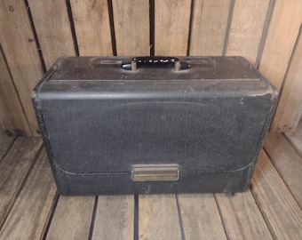 May include: A vintage black suitcase with a rectangular shape and a black handle on top. The suitcase has a metal clasp and is set against a wooden background. The suitcase appears to be made of a textured material.