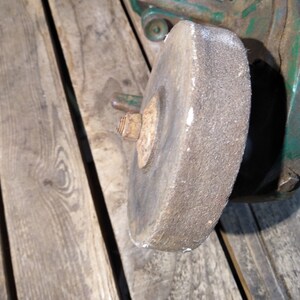 May include: Close-up of a gray grinding wheel attached to a green metal grinder. The wheel is worn and has a rough texture.