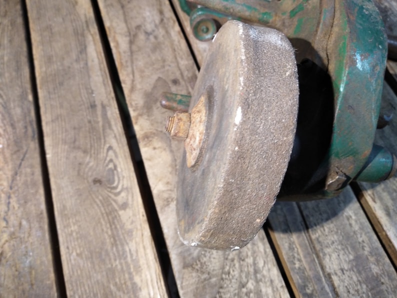May include: Close-up of a gray grinding wheel attached to a green metal grinder. The wheel is worn and has a rough texture.