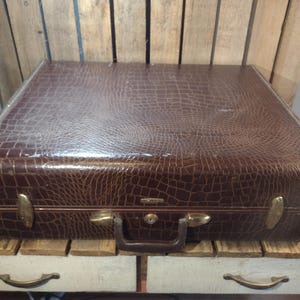 May include: A vintage brown suitcase with a faux crocodile skin texture. The suitcase has brass-colored hardware, including latches, a handle, and corner protectors. The suitcase is sitting on a wooden surface.