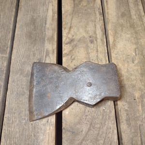 May include: A vintage, weathered axe head with a dark, metallic finish. The axe head is resting on a wooden surface. The axe head has a slightly curved blade and a rectangular shape with a small, circular rivet.