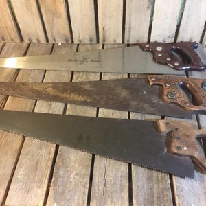 Può includere: Tre seghe a mano antiche con manici in legno e lame in metallo. La sega superiore presenta il testo "Bessy & Bess" sulla lama. Le seghe sono disposte su una superficie di legno, mostrando il loro aspetto invecchiato e la loro maestria.