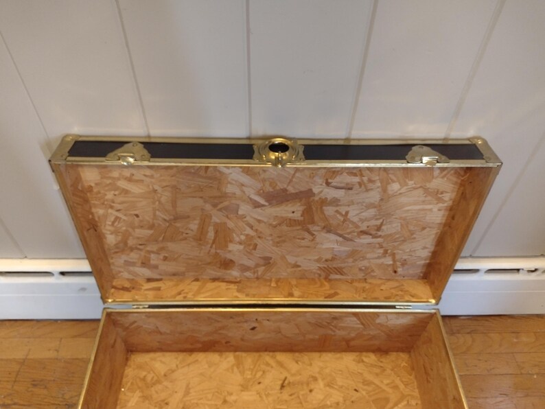 May include: An open wooden chest with a dark top and brass-colored accents. The interior is made of light-colored wood. The chest has a rectangular shape and is designed for storage. The top has a black panel with gold trim and a central lock.