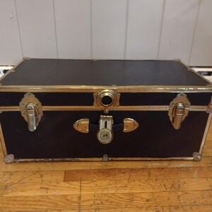 May include: A black trunk with gold-colored metal trim and hardware. The trunk has a rectangular shape with a flat top and is closed with a lock and two latches. The trunk is sitting on a wooden floor.