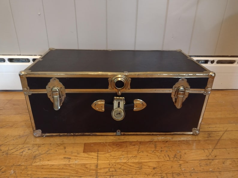 May include: A black trunk with gold-colored metal trim and hardware. The trunk has a rectangular shape with a flat top and is closed with a lock and two latches. The trunk is sitting on a wooden floor.