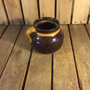 May include: A brown ceramic crock with a yellow rim and a single handle. The crock is sitting on a wooden surface.