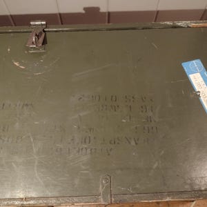 May include: A green metal box with a blue label that says "702 505 SCN". The box has several numbers and letters written on it, including "29010 55V", "88W7 91", "3.40", and "A N.Y. 1911-2021".