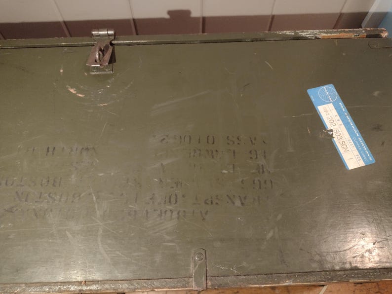 May include: A green metal box with a blue label that says "702 505 SCN". The box has several numbers and letters written on it, including "29010 55V", "88W7 91", "3.40", and "A N.Y. 1911-2021".