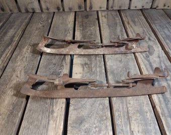 May include: Two rusty metal ice skate blades with adjustable straps. The blades are on a wooden surface.