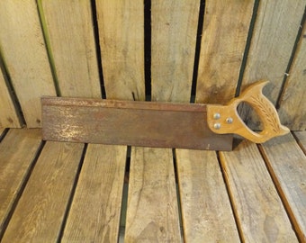 May include: A rusty metal saw with a wooden handle. The handle has a carved design of leaves. The saw is resting on a wooden surface.