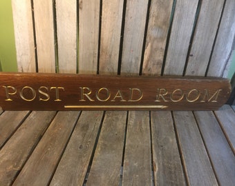 May include: A wooden sign with gold lettering that reads "POST ROAD ROOM". The sign has a gold arrow pointing to the right.