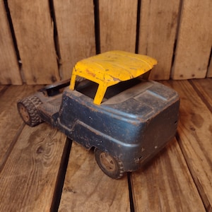 May include: A vintage toy truck with a blue body and a yellow roof. The truck has four wheels and a detailed design, showing signs of wear. The toy is made of metal and sits on a wooden surface.