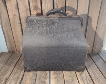 May include: A vintage black leather doctor's bag with a textured surface and a curved handle. The bag is rectangular with rounded corners and sits on a wooden surface. The bag has metal clasps and hinges.