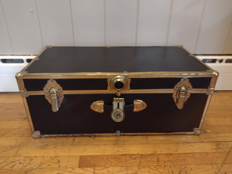 May include: A vintage black trunk with gold-colored metal trim and hardware. The trunk has a rectangular shape with a flat top and is secured with a lock and latches. The trunk is sitting on a wooden floor.