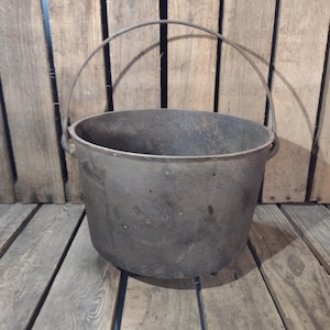 May include: A dark gray cast iron pot with a curved metal handle. The pot is round with a wide opening and sits on a wooden surface. The pot is likely used for cooking or storage.