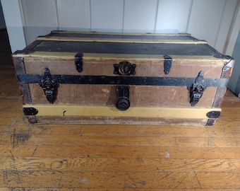 May include: A vintage wooden trunk with black metal hardware and a yellow painted stripe. The trunk has a worn, distressed look and is sitting on a wooden floor.