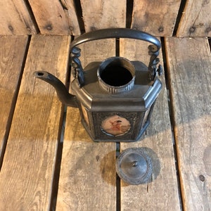 May include: A black metal teapot with a square body and a lid. The teapot has a handle with decorative scrolls and a spout. The teapot is decorated with a painted scene of a person in a landscape. The teapot is sitting on a wooden surface.
