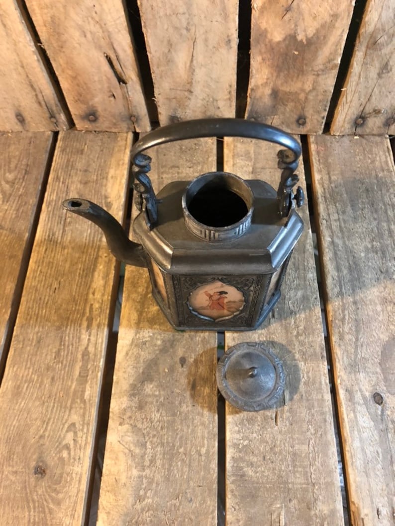 May include: A black metal teapot with a square body and a lid. The teapot has a handle with decorative scrolls and a spout. The teapot is decorated with a painted scene of a person in a landscape. The teapot is sitting on a wooden surface.