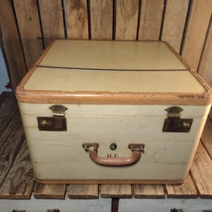 May include: Vintage cream-colored suitcase with tan leather trim and brass hardware. The suitcase has a rectangular shape with a curved top and a handle with the initials "H.E." on it. The suitcase is sitting on a wooden surface.