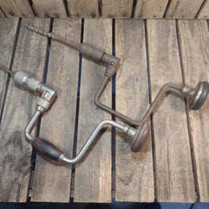 May include: Two antique hand drills with wooden handles and metal components. The drills are lying on a weathered wooden surface. One drill has a dark brown handle and a silver metal body. The other drill has a brown handle and a silver metal body.