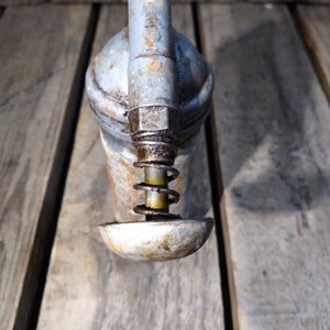 May include: A vintage metal oil can with a spring-loaded pump and a gray finish. The can has a worn, rustic look and is sitting on a wooden surface.