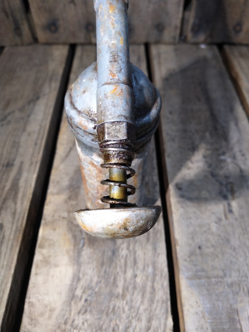 May include: A vintage metal oil can with a spring-loaded pump and a gray finish. The can has a worn, rustic look and is sitting on a wooden surface.