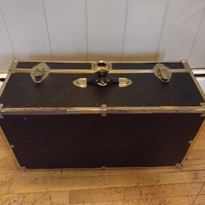 May include: A black trunk with gold-colored metal accents. The trunk has a rectangular shape with a hinged lid and metal clasps. The corners and edges are reinforced with gold-colored metal. The trunk is sitting on a wooden floor.