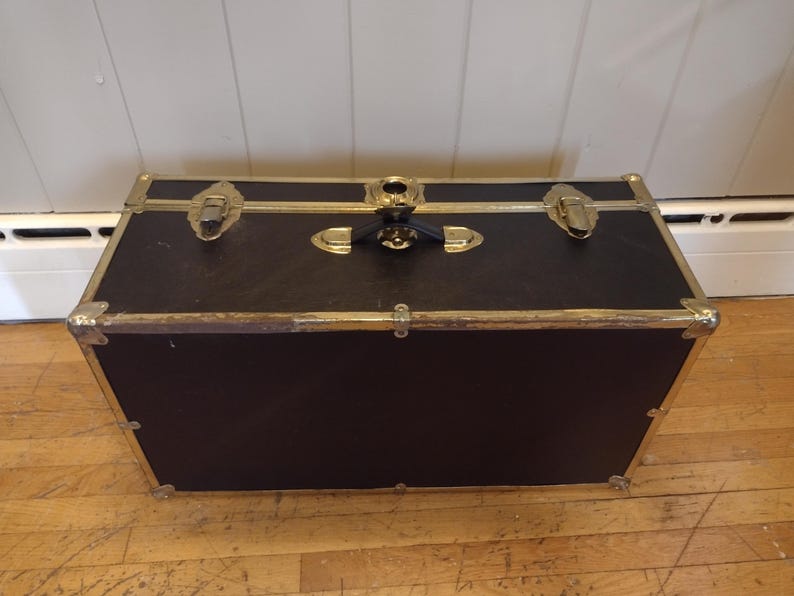 May include: A black trunk with gold-colored metal accents. The trunk has a rectangular shape with a hinged lid and metal clasps. The corners and edges are reinforced with gold-colored metal. The trunk is sitting on a wooden floor.