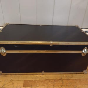 May include: A rectangular black trunk with gold-colored metal trim and corner protectors. The trunk has a hinged lid and metal latches. The trunk is sitting on a wooden floor.