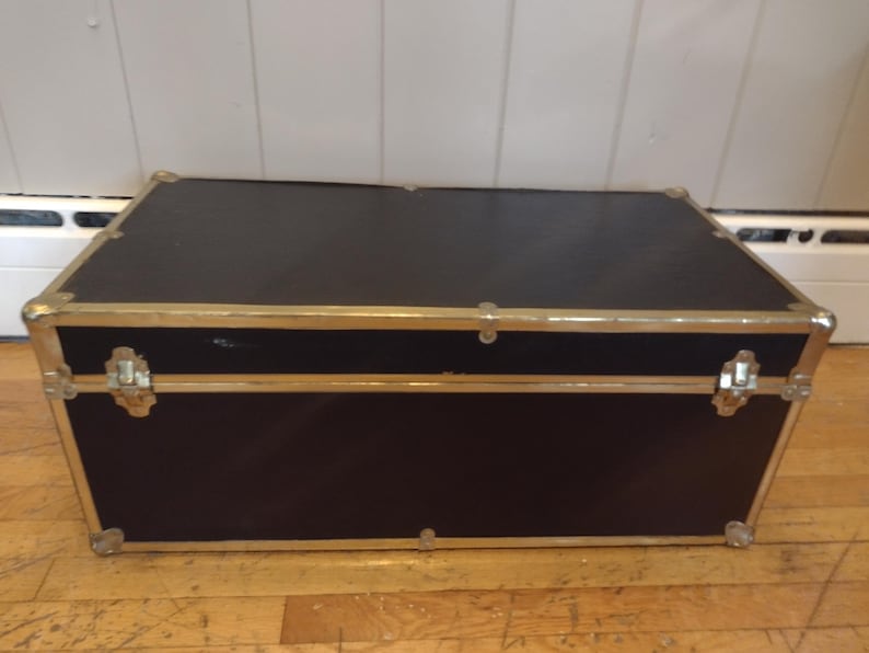 May include: A rectangular black trunk with gold-colored metal trim and corner protectors. The trunk has a hinged lid and metal latches. The trunk is sitting on a wooden floor.