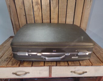 May include: Vintage olive green suitcase with silver-toned hardware, handle, and trim. The suitcase is rectangular and sits on a wooden table with a drawer. The suitcase is a classic travel accessory.