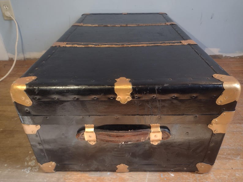 Puede incluir: Ba&uacute;l de equipaje vintage negro con detalles dorados y asa de cuero.