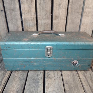 May include: Vintage teal metal toolbox with a handle and a latch. The toolbox has a weathered appearance with rust spots and horizontal ridges. A small emblem is visible on the front.