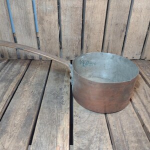 May include: A copper saucepan with a long handle. The pot has a rounded shape and a tarnished finish, suggesting age. The handle extends from the side, curving upwards. The pan sits on a wooden surface.