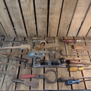 May include: A collection of vintage woodworking tools, including planes, saws, screwdrivers, and clamps, arranged on a wooden surface. The tools show signs of age and use, with various handle styles and blade shapes. The tools are in various sizes.