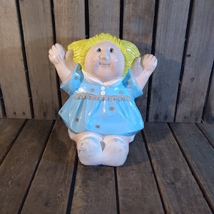 May include: A ceramic figurine of a girl with blonde hair, wearing a blue dress with white buttons and a white collar. She has a big smile and is holding her arms up in the air.