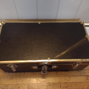 May include: A rectangular black trunk with gold-colored metal trim and corner protectors. The trunk has a latch closure and is sitting on a wooden floor. The trunk appears to be vintage or antique.