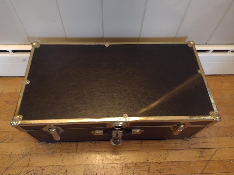 May include: A rectangular black trunk with gold-colored metal trim and corner protectors. The trunk has a latch closure and is sitting on a wooden floor. The trunk appears to be vintage or antique.