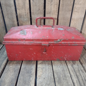 May include: A vintage red metal toolbox with a handle and latch. The toolbox shows signs of wear, with paint chipping and a distressed appearance. The rectangular box is set against a wooden background.