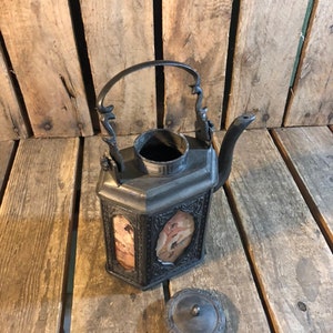 May include: A black metal teapot with a decorative handle and a lid. The teapot has a square body with a rectangular opening on each side, revealing a patterned design. The teapot is sitting on a wooden surface.