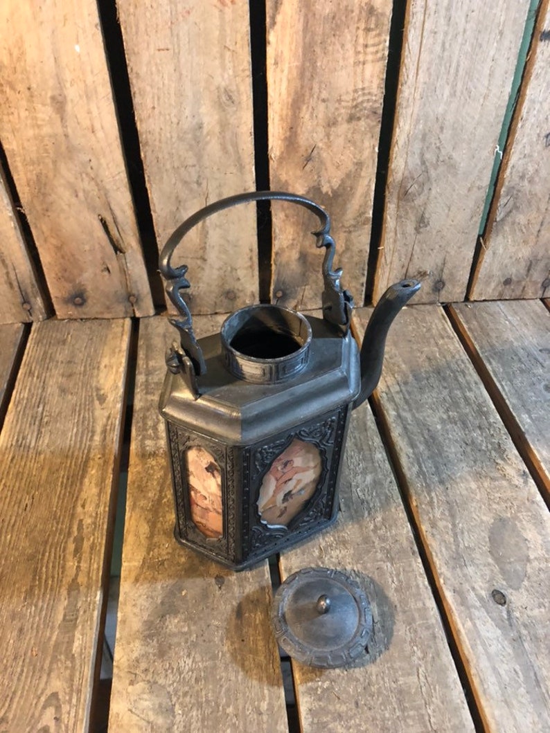 May include: A black metal teapot with a decorative handle and a lid. The teapot has a square body with a rectangular opening on each side, revealing a patterned design. The teapot is sitting on a wooden surface.