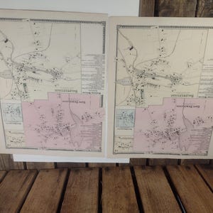 May include: Two antique maps of Baldwinsville, featuring detailed town layouts and surrounding areas. The maps have a cream-colored background with black and pink details. The maps are displayed on a wooden surface.