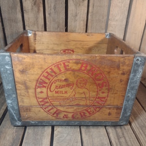 May include: Vintage wooden milk crate with metal corner reinforcements. The crate features a red circular logo that reads "WHITE BROS MILK & CREAM" with a graphic of a baby. The crate has hand-cut holes on the sides.