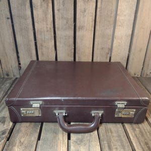 May include: A brown leather briefcase with a handle and combination locks. The vintage case is rectangular and sits on a wooden surface. The briefcase has metal hardware and a stitched design.