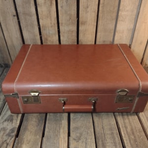 May include: A vintage brown suitcase with brass hardware, including latches and a handle. The suitcase is rectangular with rounded corners and white stitching along the edges. It sits on a wooden surface.
