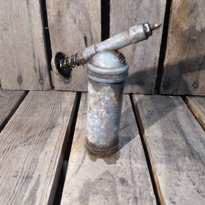 May include: A vintage, silver-colored grease gun with a black rubber tip and a metal spring. The gun is sitting on a wooden surface.