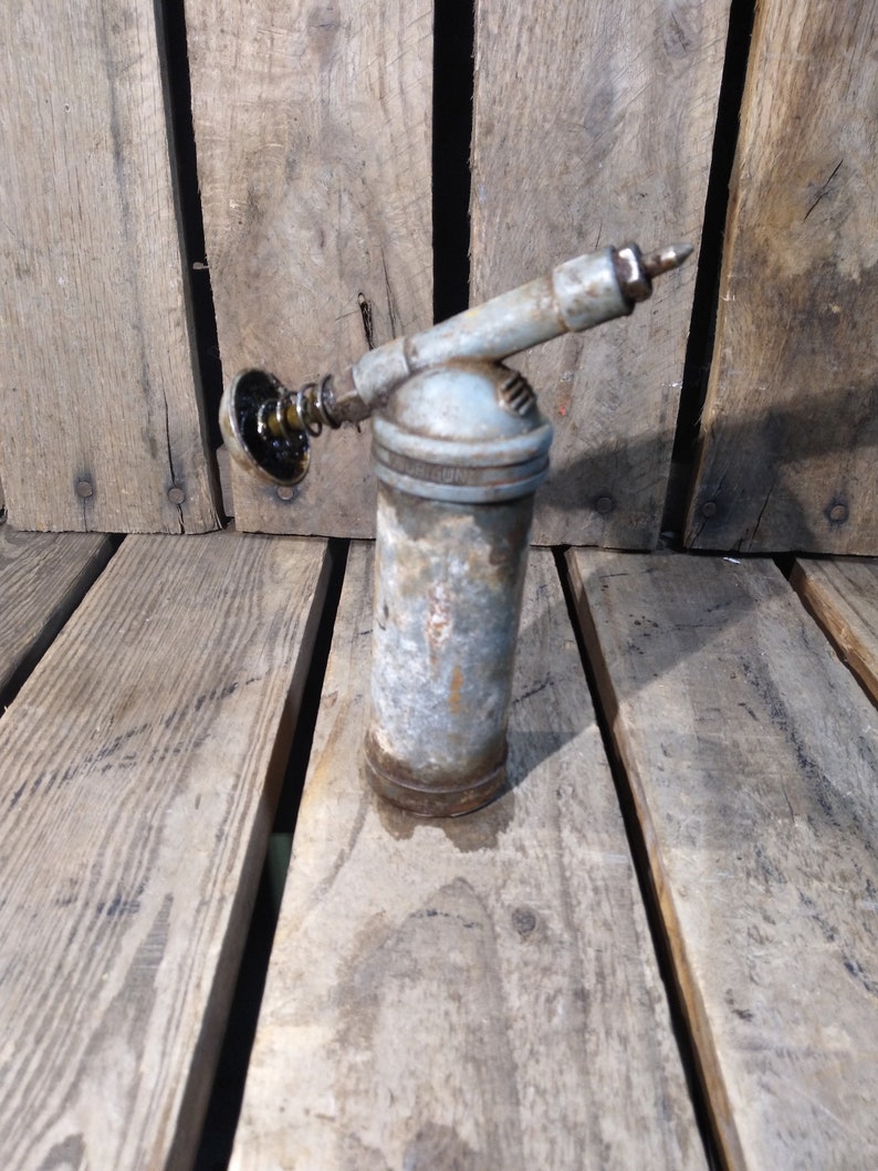 May include: A vintage, silver-colored grease gun with a black rubber tip and a metal spring. The gun is sitting on a wooden surface.
